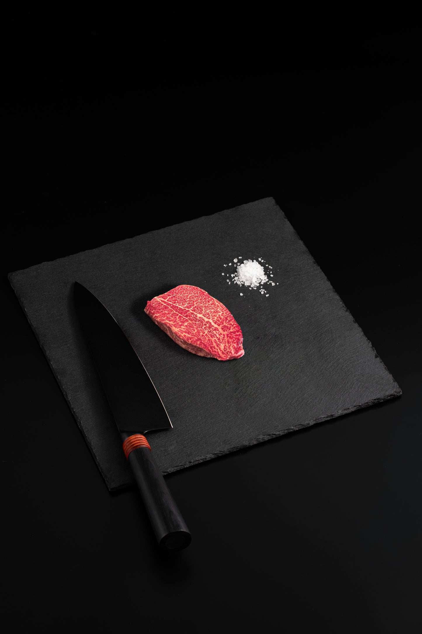 Single marbled Wagyu steak with sea salt and black chef’s knife on a dark slate board