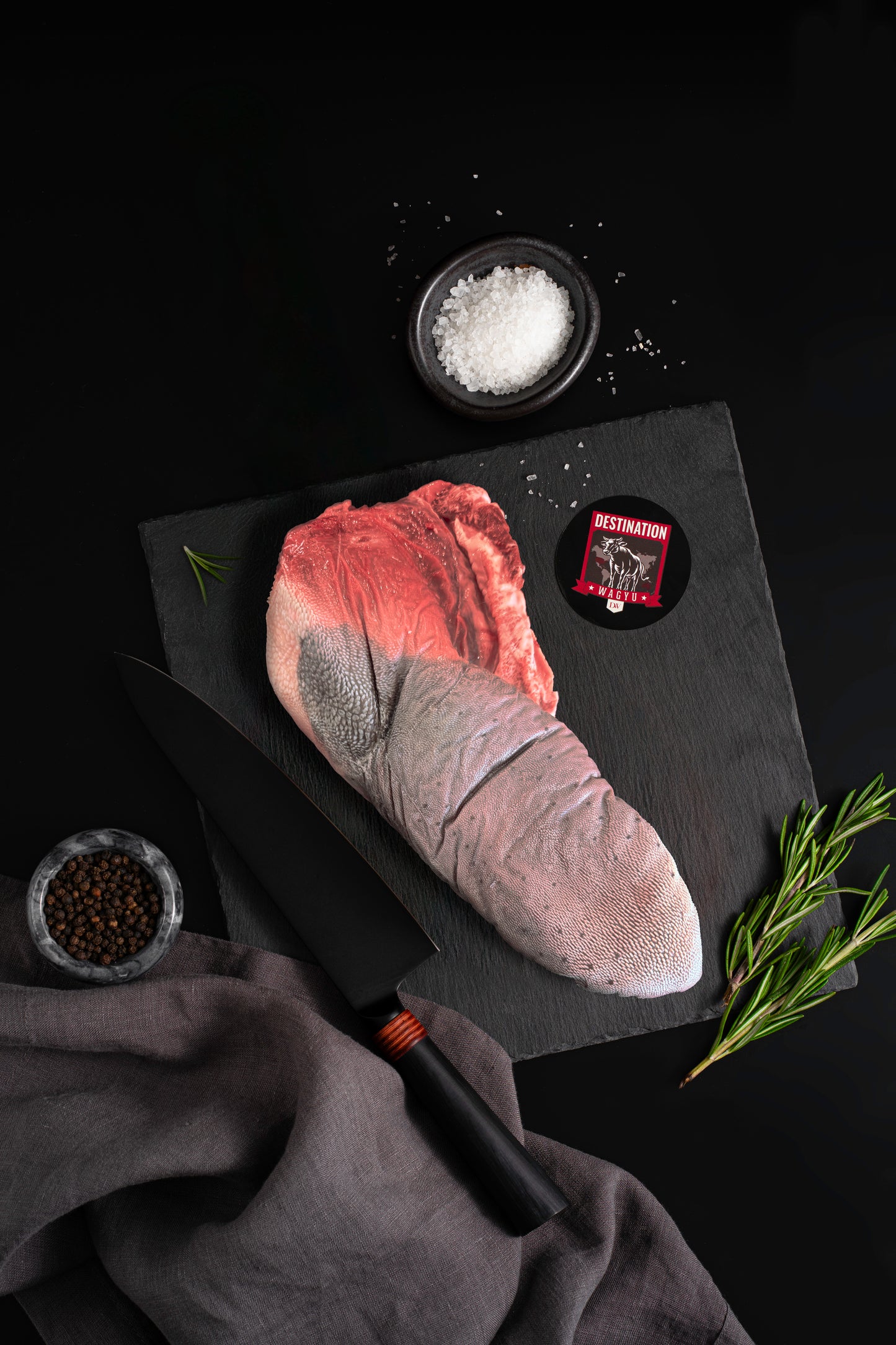 Australian Wagyu beef tongue on a black slate board with Destination Wagyu label, sea salt bowl, peppercorns, and rosemary on a dark background