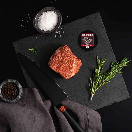 A Japanese A5 Wagyu Top Sirloin rests on a slate board with rosemary, a bowl of salt, a bowl of pepper, and a black knife.