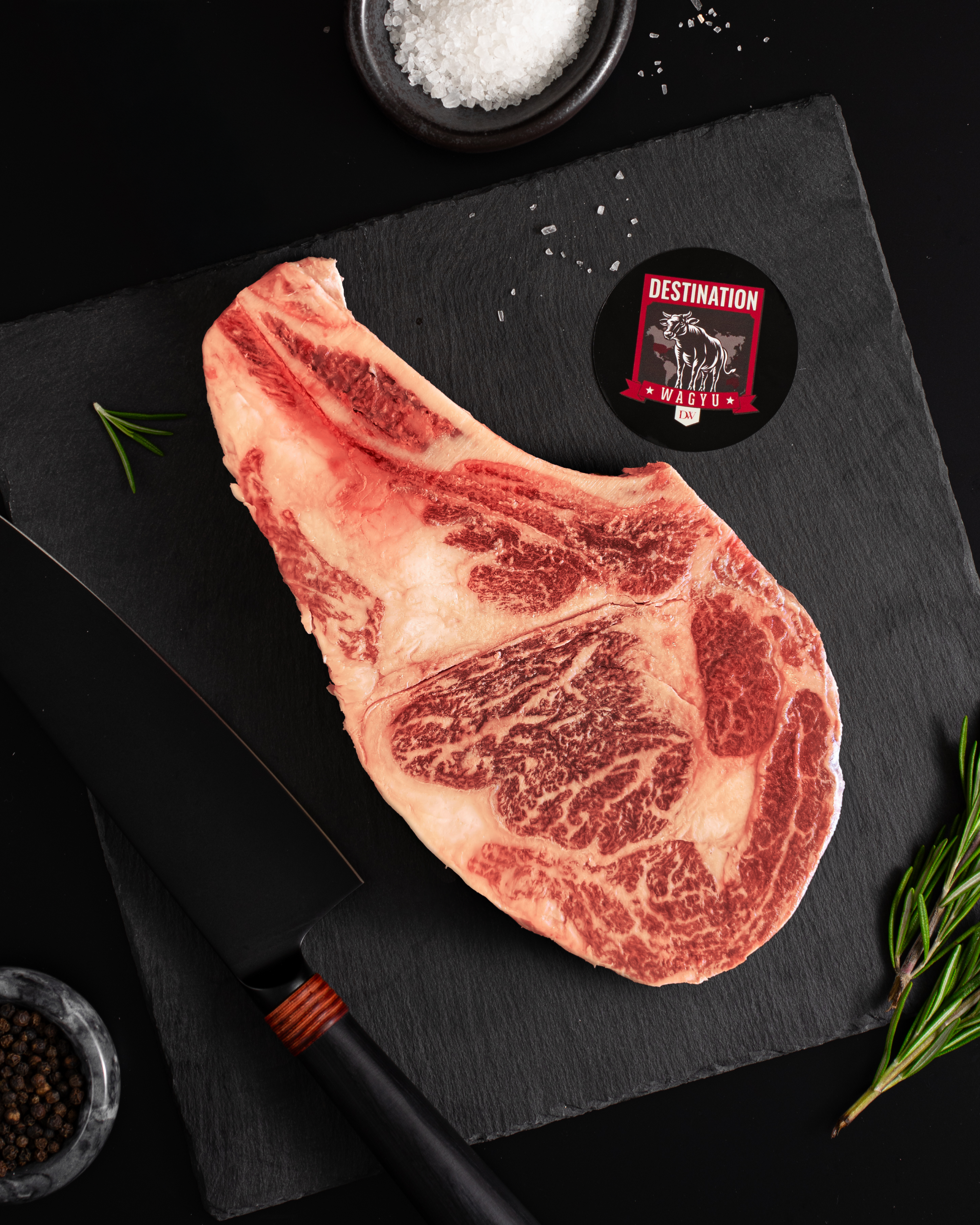Australian Wagyu bone-in ribeye steak (18oz, Stone Axe) on a dark slate board with Destination Wagyu logo, chef’s knife, sea salt and rosemary garnish.