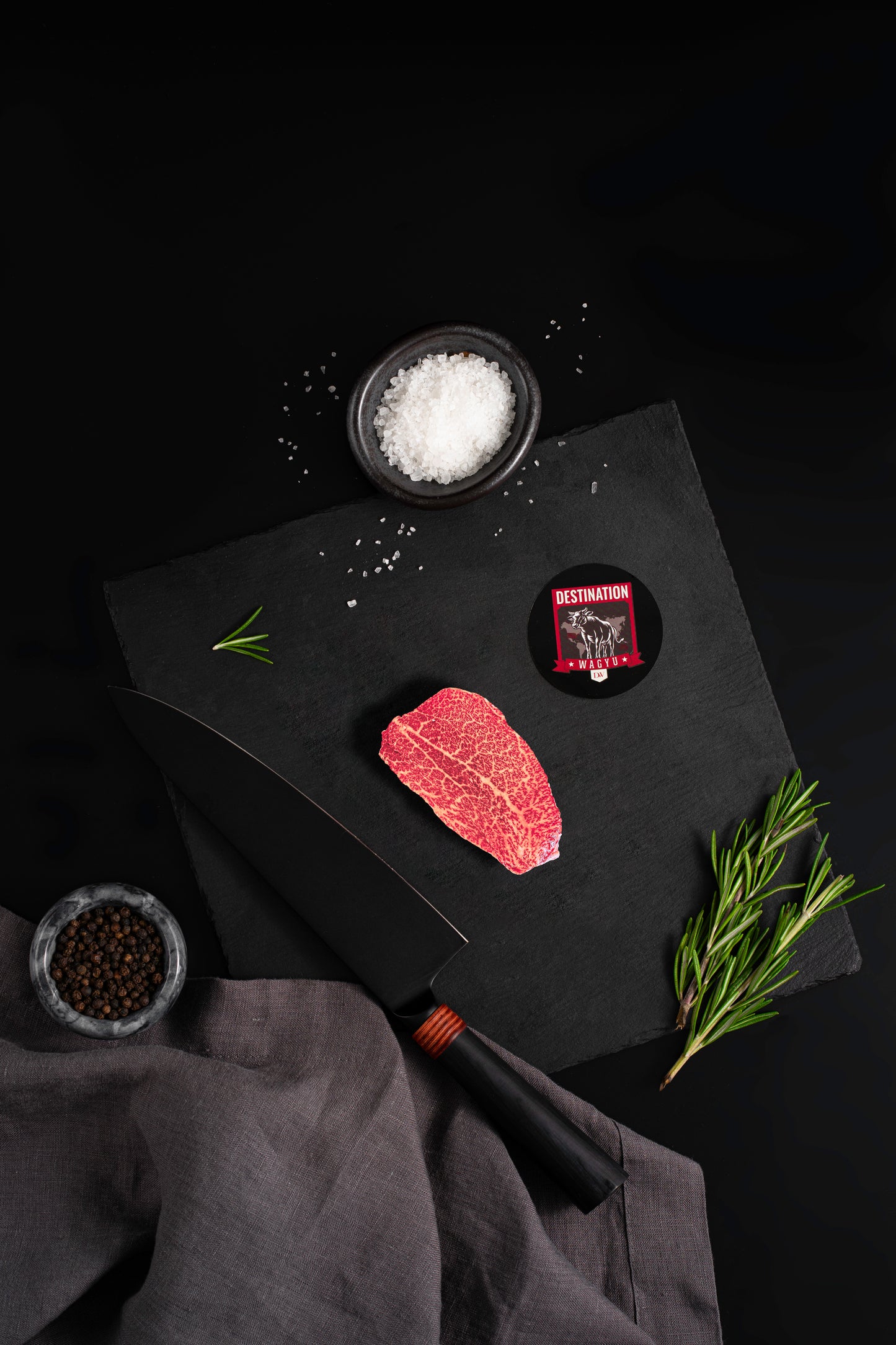 Marbled Wagyu steak on slate board with sea salt, rosemary, peppercorns, black chef’s knife, and Destination Wagyu logo on a dark background