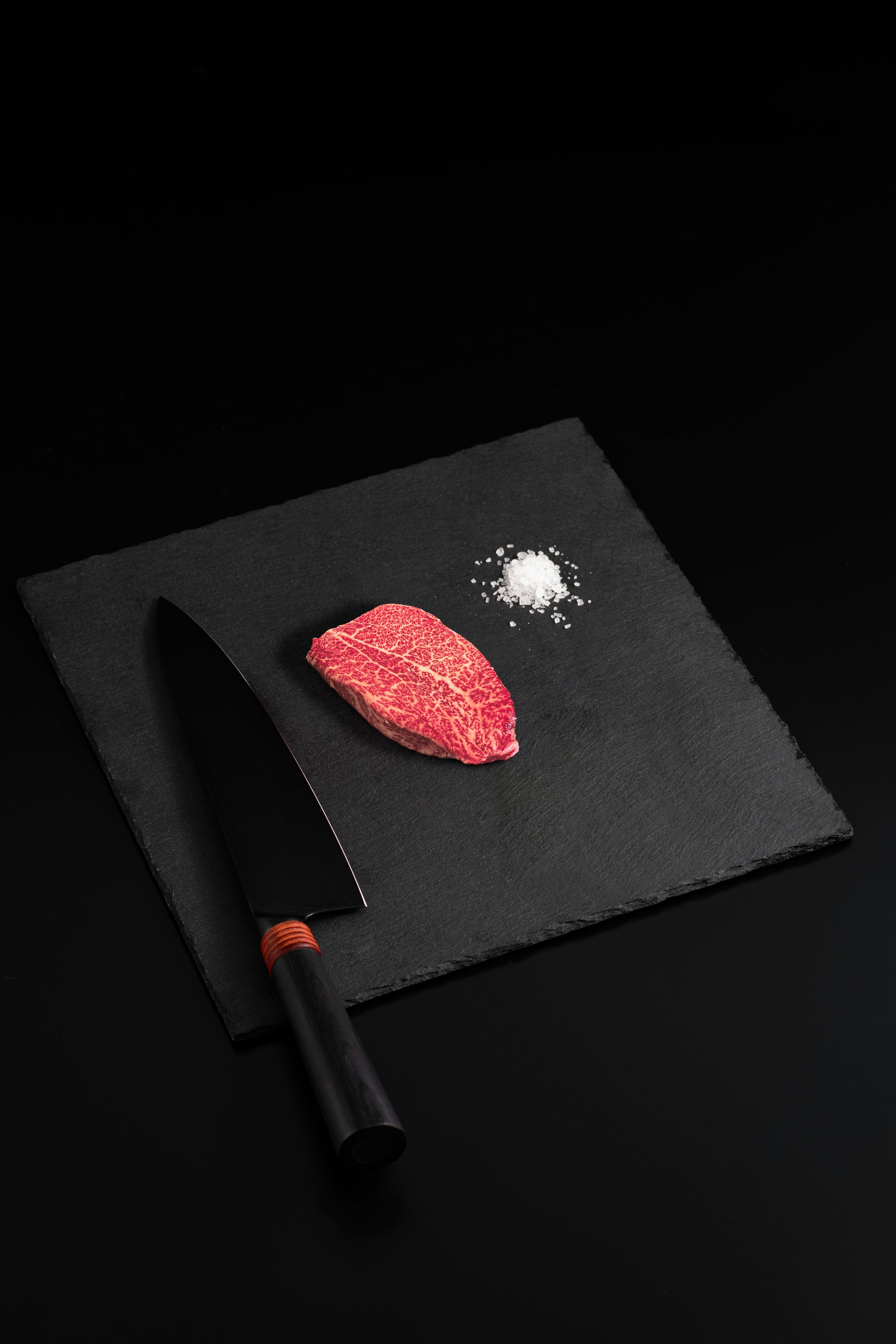 Single marbled Wagyu steak with sea salt and black chef’s knife on a dark slate board