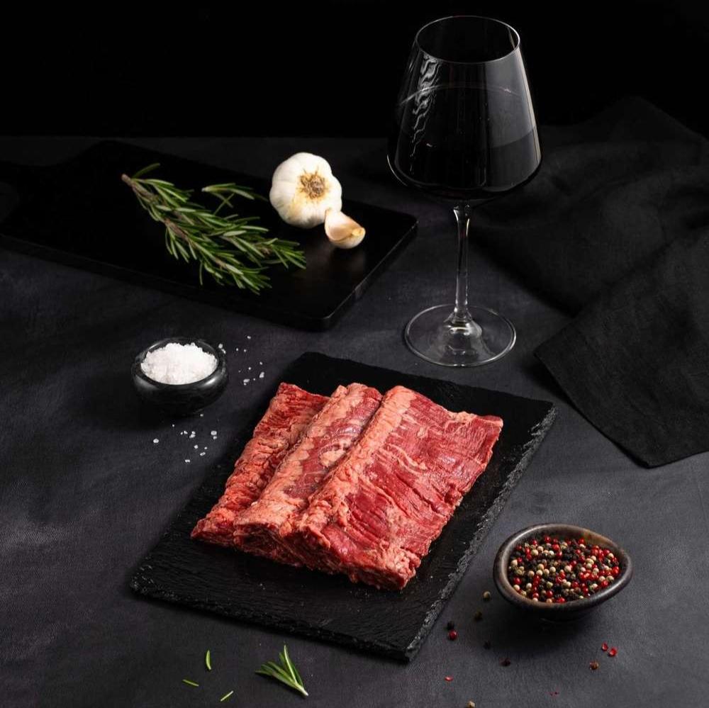 Raw Australian Wagyu Outside Skirt Steak rests on a black slate surrounded by salt, pepper, rosemary, garlic, and complemented by a glass of red wine on a dark surface.