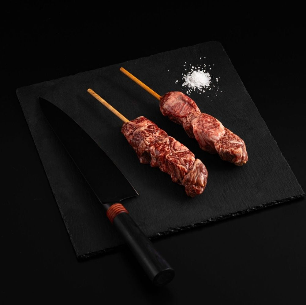 Two Japanese A5 Wagyu Filet Mignon Skewers with salt and chef's knife on a black slate, isolated on black background.