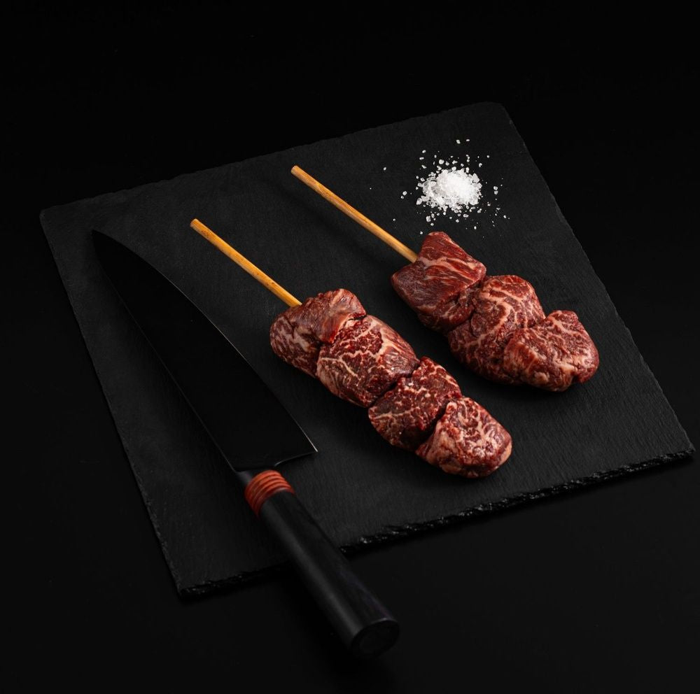 Two Australian Wagyu Filet Mignon Skewers with salt and chef's knife on a black slate, isolated on black background.