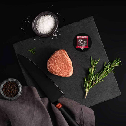 A Japanese A5 Wagyu Miyazaki Filet Steak rests on a slate board with rosemary, a bowl of salt, a bowl of pepper, and a black knife.