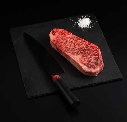 An Australian Wagyu New York Striploin Steak with salt and chef's knife on a black slate, isolated on black background.