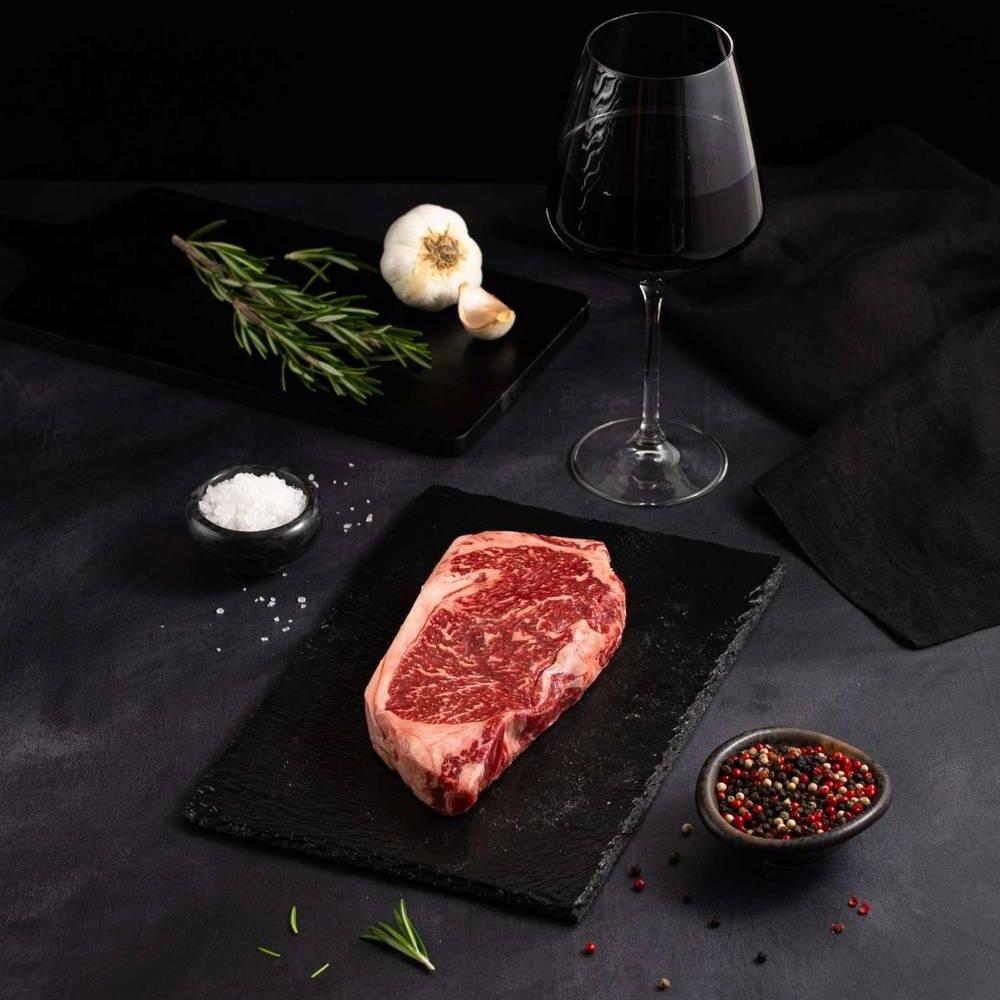 A Raw Australian Wagyu New York Striploin Steak rests on a black slate surrounded by salt, pepper, rosemary, garlic, and complemented by a glass of red wine on a dark surface.