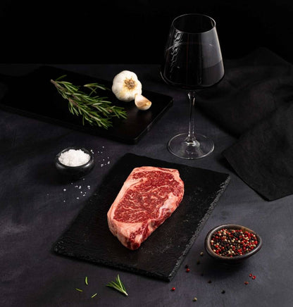 A succulent American Wagyu New York Masami Steak, raw and marbled with BMS 8-9 perfection, rests on a black slate surrounded by salt, pepper, rosemary, garlic, and complemented by a glass of red wine on a dark surface.