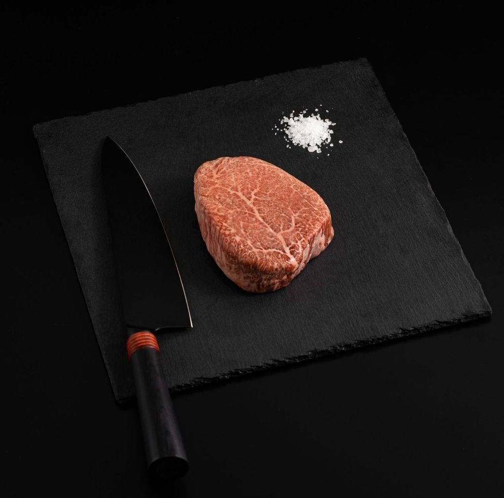 A Japanese A5 Wagyu Kobe Wine Filet Steak with salt and chef's knife on a black slate, isolated on black background.