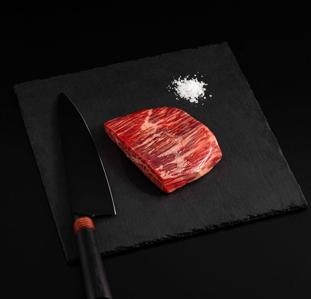 An Australian Wagyu Flank Steak with salt and chef's knife on a black slate, isolated on black background.