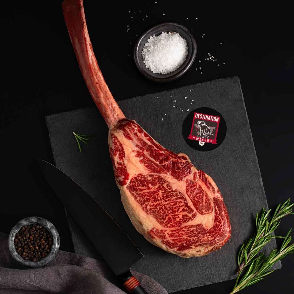 An Australian Wagyu Tomahawk Steak rests on a slate board with rosemary, a bowl of salt, a bowl of pepper, and a black knife.