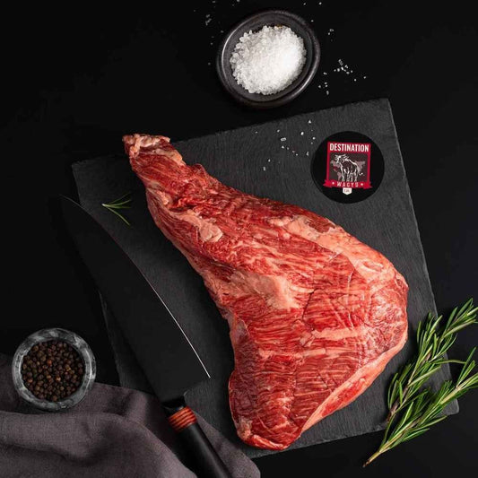 An American Wagyu Masami Tri Tip rests on a slate board with rosemary, a bowl of salt, a bowl of pepper, and a black knife.