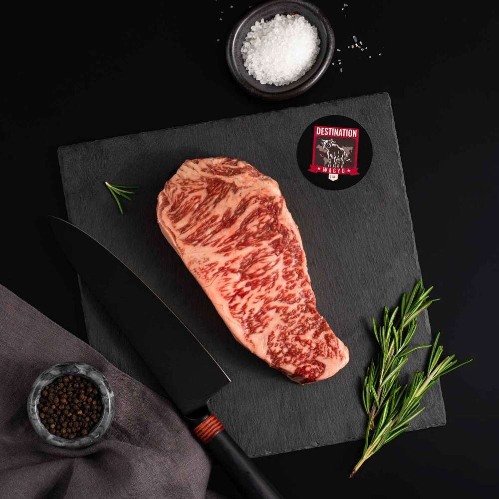 An Australian Wagyu Full Blood New York Striploin Steak rests on a slate board with rosemary, a bowl of salt, a bowl of pepper, and a black knife.