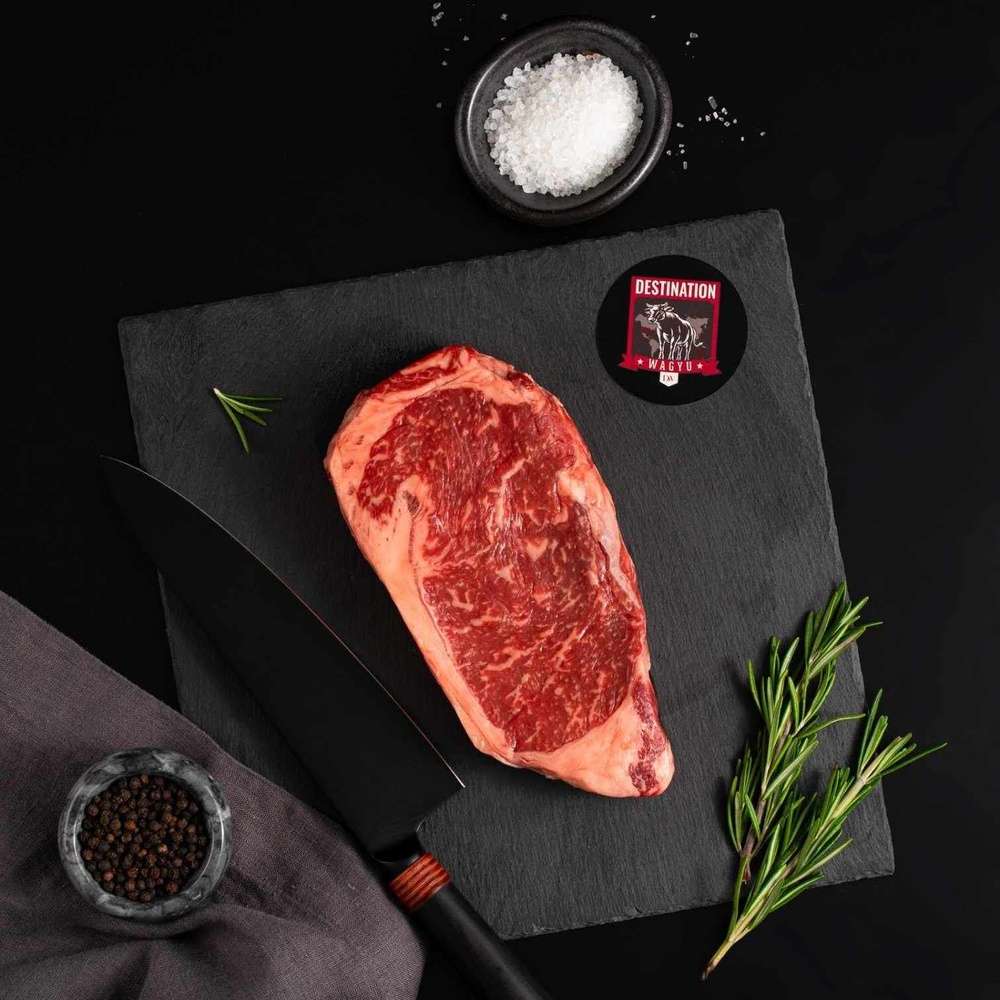 An Australian Wagyu New York Striploin Steak rests on a slate board with rosemary, a bowl of salt, a bowl of pepper, and a black knife.