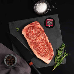 A Japanese A5 Wagyu Kobe Wine New York Strip Steak rests on a slate board with rosemary, a bowl of salt, a bowl of pepper, and a black knife.