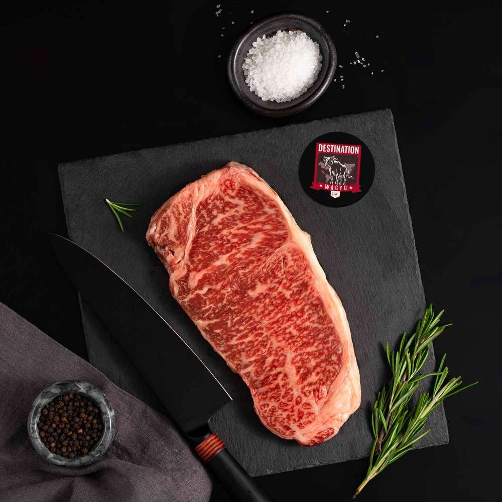 A Japanese A5 Wagyu Striploin Miyazaki Steak rests on a slate board with rosemary, a bowl of salt, a bowl of pepper, and a black knife.