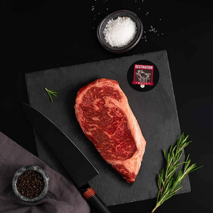 A 14-16 oz American Wagyu New York Masami Steak rests on a slate board with rosemary, a bowl of salt, a bowl of pepper, and a black knife.