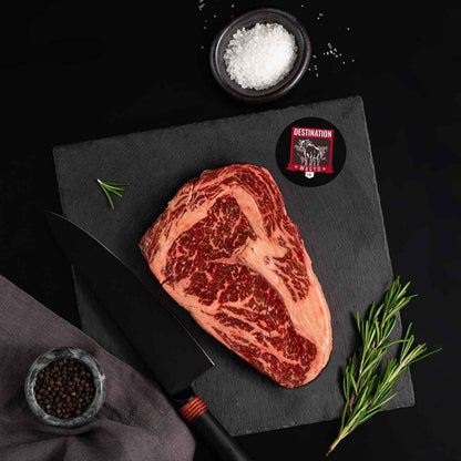 An Australian Wagyu Jade Full Blood Ribeye Steak rests on a slate board with rosemary, a bowl of salt, a bowl of pepper, and a black knife.