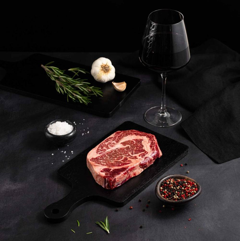 A Raw Australian Wagyu Ribeye Steak rests on a black slate surrounded by salt, pepper, rosemary, garlic, and complemented by a glass of red wine on a dark surface.