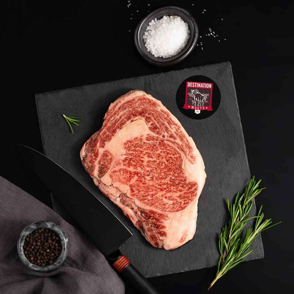 A Japanese A5 Wagyu Miyazaki Ribeye Steak rests on a slate board with rosemary, a bowl of salt, a bowl of pepper, and a black knife.