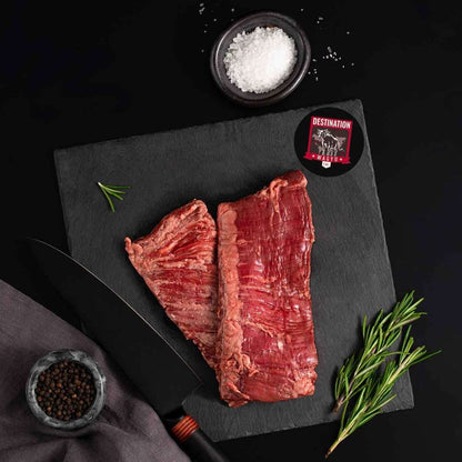 Australian Wagyu Outside Skirt Steak rests on a slate board with rosemary, a bowl of salt, a bowl of pepper, and a black knife.