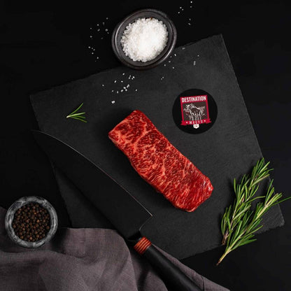 An Australian Wagyu Full Blood Denver Steak rests on a slate board with rosemary, a bowl of salt, a bowl of pepper, and a black knife.