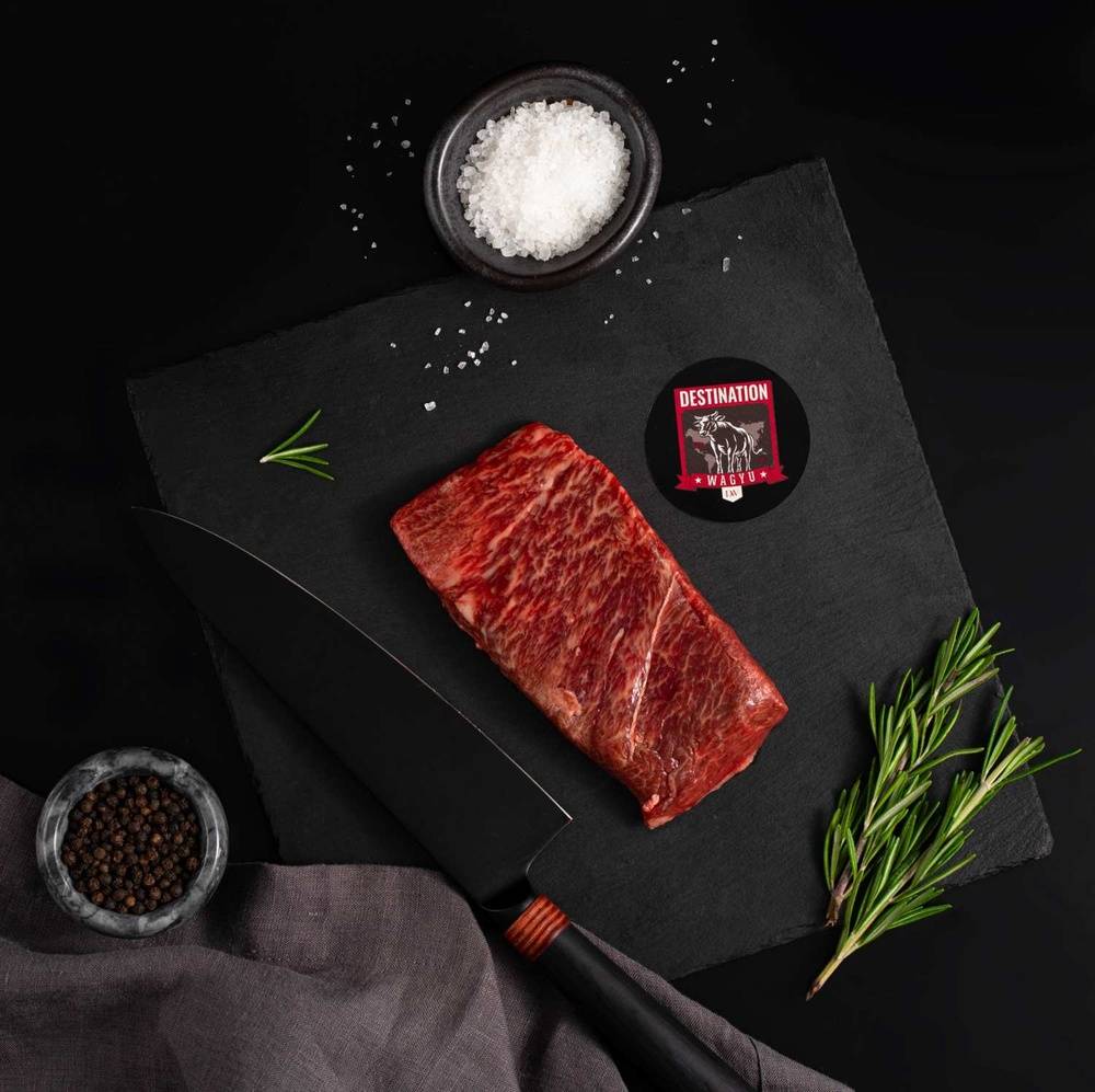 An Australian Wagyu Full Blood Flat Iron Steak rests on a slate board with rosemary, a bowl of salt, a bowl of pepper, and a black knife.