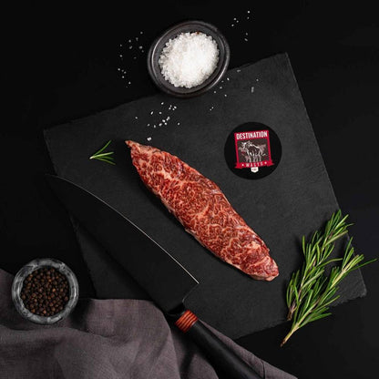 A Japanese A5 Wagyu Flap Meat Steak rests on a slate board with rosemary, a bowl of salt, a bowl of pepper, and a black knife.