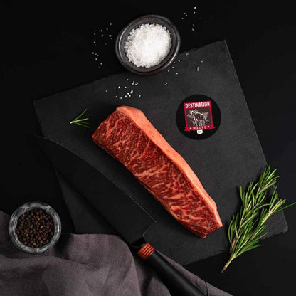 An Australian Wagyu Top Coulotte Picanha Steak rests on a slate board with rosemary, a bowl of salt, a bowl of pepper, and a black knife.