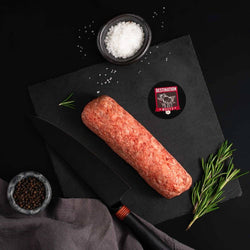 A slab of Japanese A5 Wagyu Ground Beef rests on a slate board with rosemary, a bowl of salt, a bowl of pepper, and a black knife.