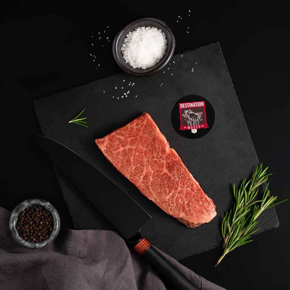 A Japanese A5 Wagyu Denver Steak rests on a slate board with rosemary, a bowl of salt, a bowl of pepper, and a black knife.