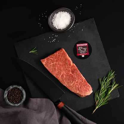 A Japanese A5 Wagyu Denver Steak rests on a slate board with rosemary, a bowl of salt, a bowl of pepper, and a black knife.