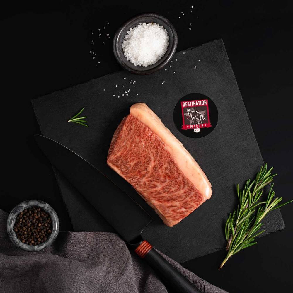 A Japanese A5 Top Coulotte Wagyu Picanha Steak rests on a slate board with rosemary, a bowl of salt, a bowl of pepper, and a black knife.