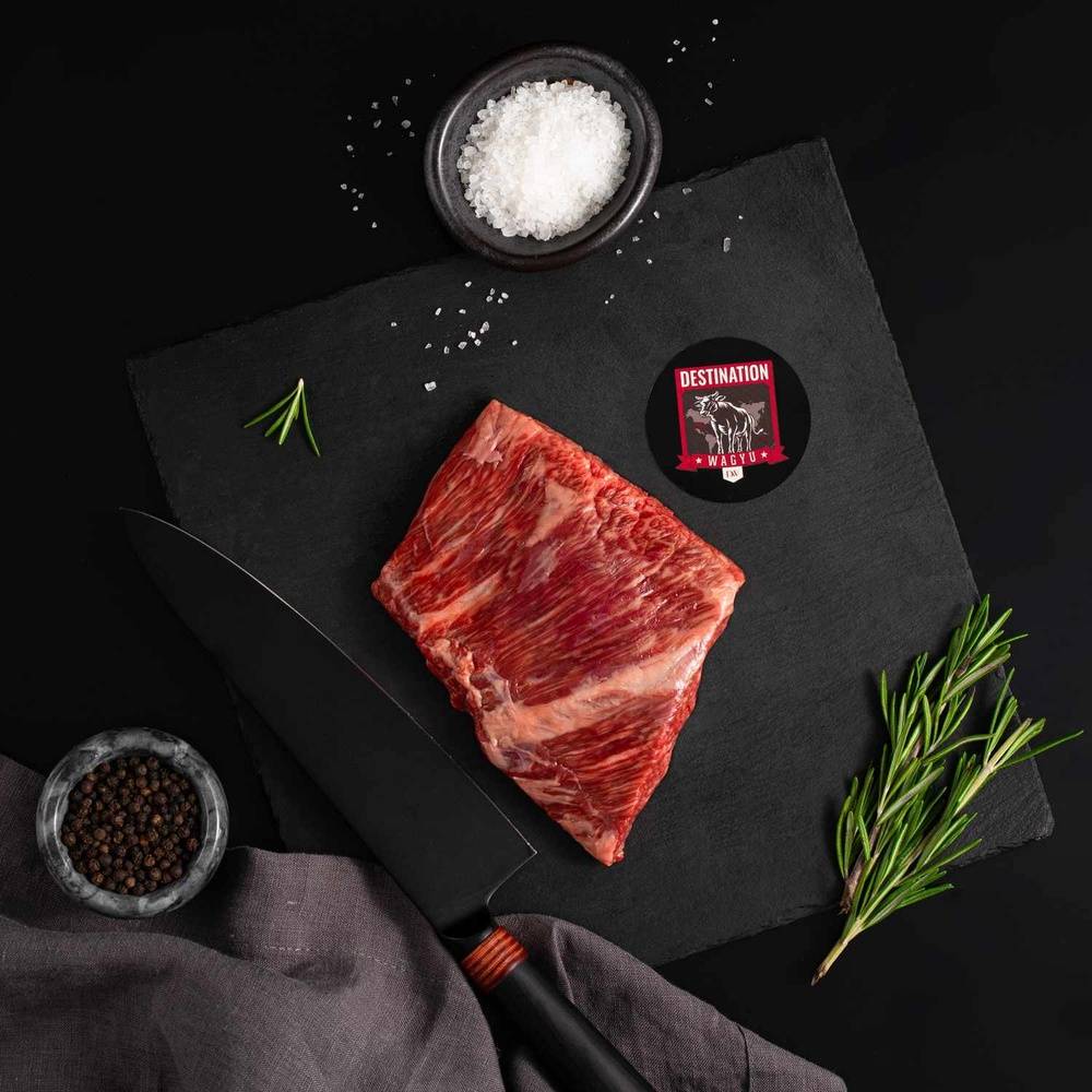 A Japanese A5 Wagyu Rib Cap Steak rests on a slate board with rosemary, a bowl of salt, a bowl of pepper, and a black knife.