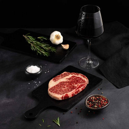 A Raw Australian Wagyu Tomahawk Ribeye Steak rests on a black slate surrounded by salt, pepper, rosemary, garlic, and complemented by a glass of red wine on a dark surface.