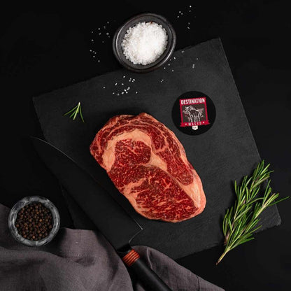 An Australian Wagyu Ribeye Steak rests on a slate board with rosemary, a bowl of salt, a bowl of pepper, and a black knife.