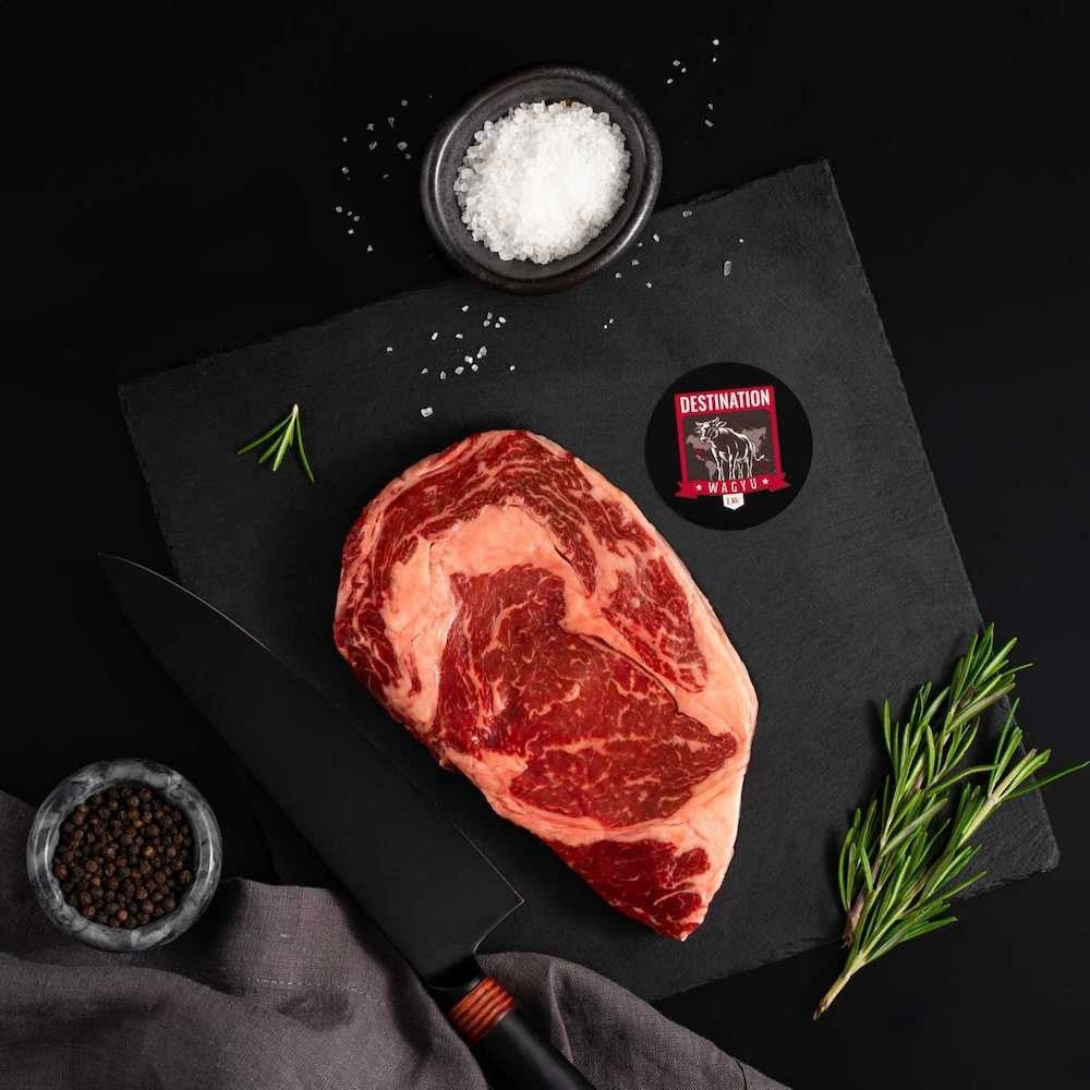 An American Wagyu Masami Ribeye Steak rests on a slate board with rosemary, a bowl of salt, a bowl of pepper, and a black knife.