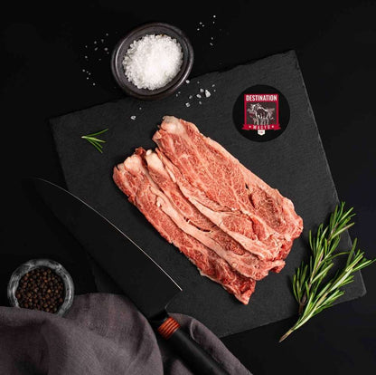 A Japanese A5 Wagyu Sliced Chuck Roll rests on a slate board with rosemary, a bowl of salt, a bowl of pepper, and a black knife.