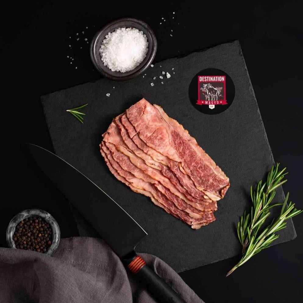 An Australian Wagyu Beef Bacon rests on a slate board with rosemary, a bowl of salt, a bowl of pepper, and a black knife.