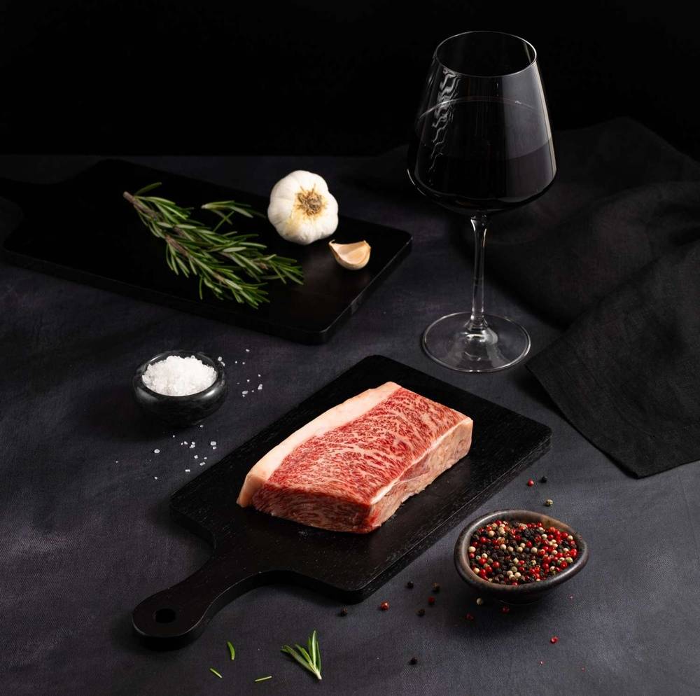 A Raw Japanese A5 Top Coulotte Wagyu Picanha Steak rests on a black slate surrounded by salt, pepper, rosemary, garlic, and complemented by a glass of red wine on a dark surface.