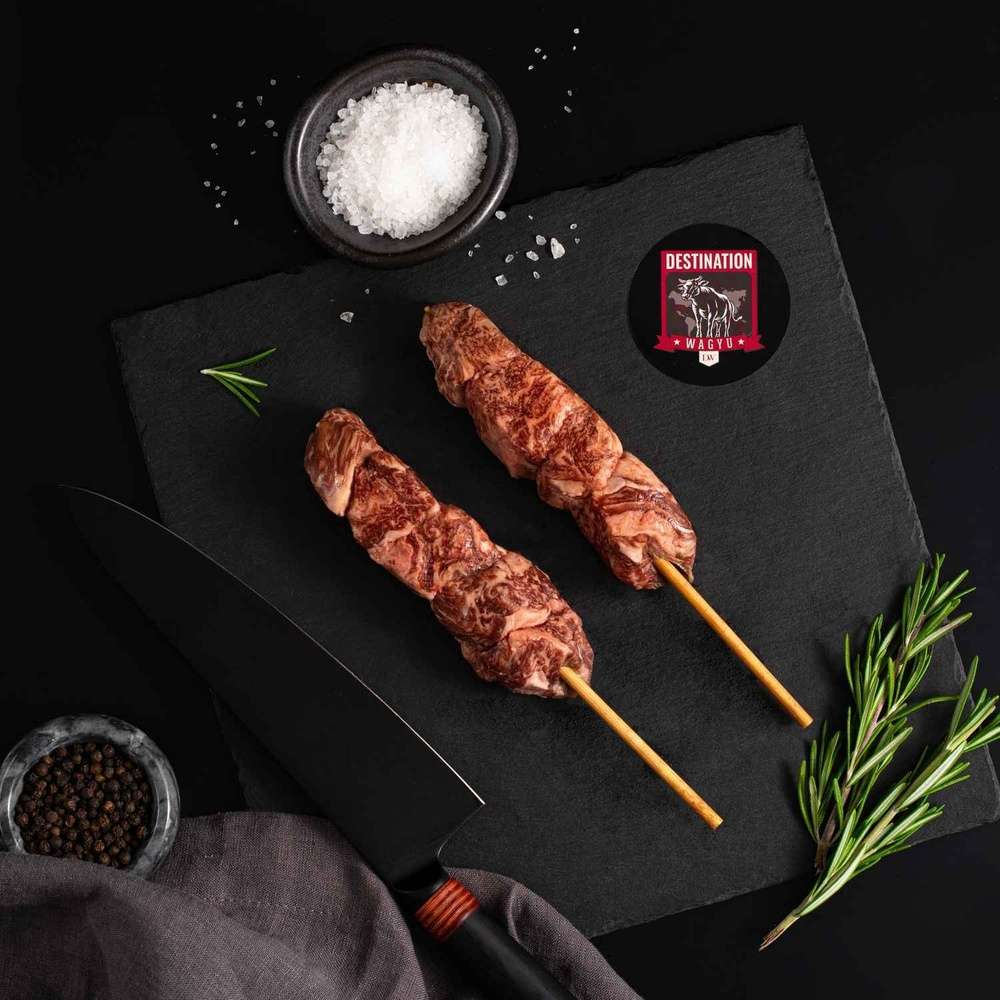 Two Japanese A5 Wagyu Filet Mignon Skewers rests on a slate board with rosemary, a bowl of salt, a bowl of pepper, and a black knife.