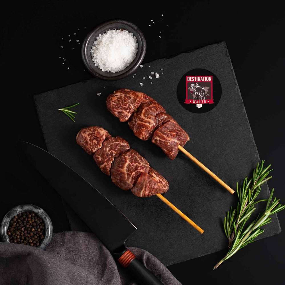 Two Australian Wagyu Filet Mignon Skewers rests on a slate board with rosemary, a bowl of salt, a bowl of pepper, and a black knife.