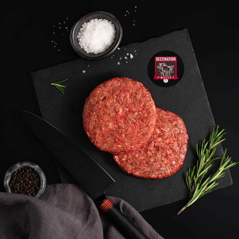 Two Australian Wagyu Beef Patties rests on a slate board with rosemary, a bowl of salt, a bowl of pepper, and a black knife.