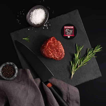 An Australian Wagyu Fullblood Jade Filet Mignon rests on a slate board with rosemary, a bowl of salt, a bowl of pepper, and a black knife.