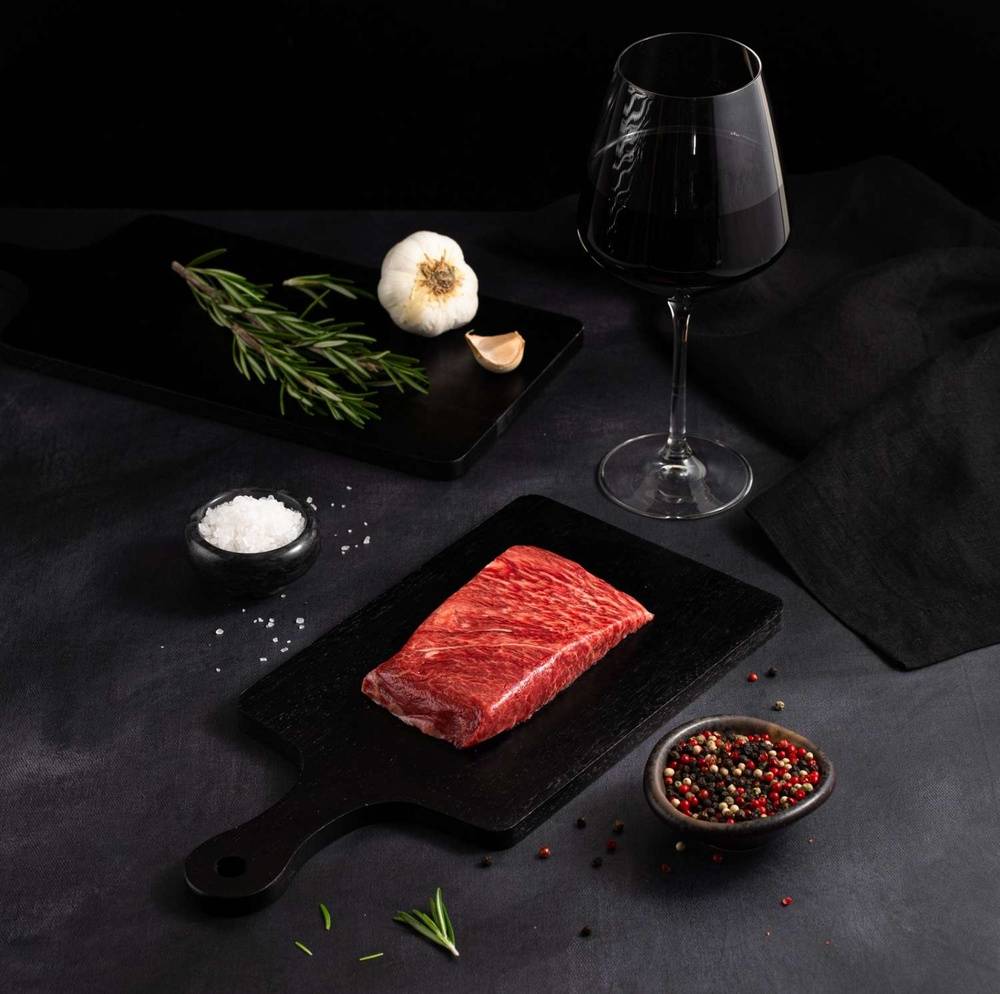 A Raw Australian Wagyu Full Blood Flat Iron Steak rests on a black slate surrounded by salt, pepper, rosemary, garlic, and complemented by a glass of red wine on a dark surface.