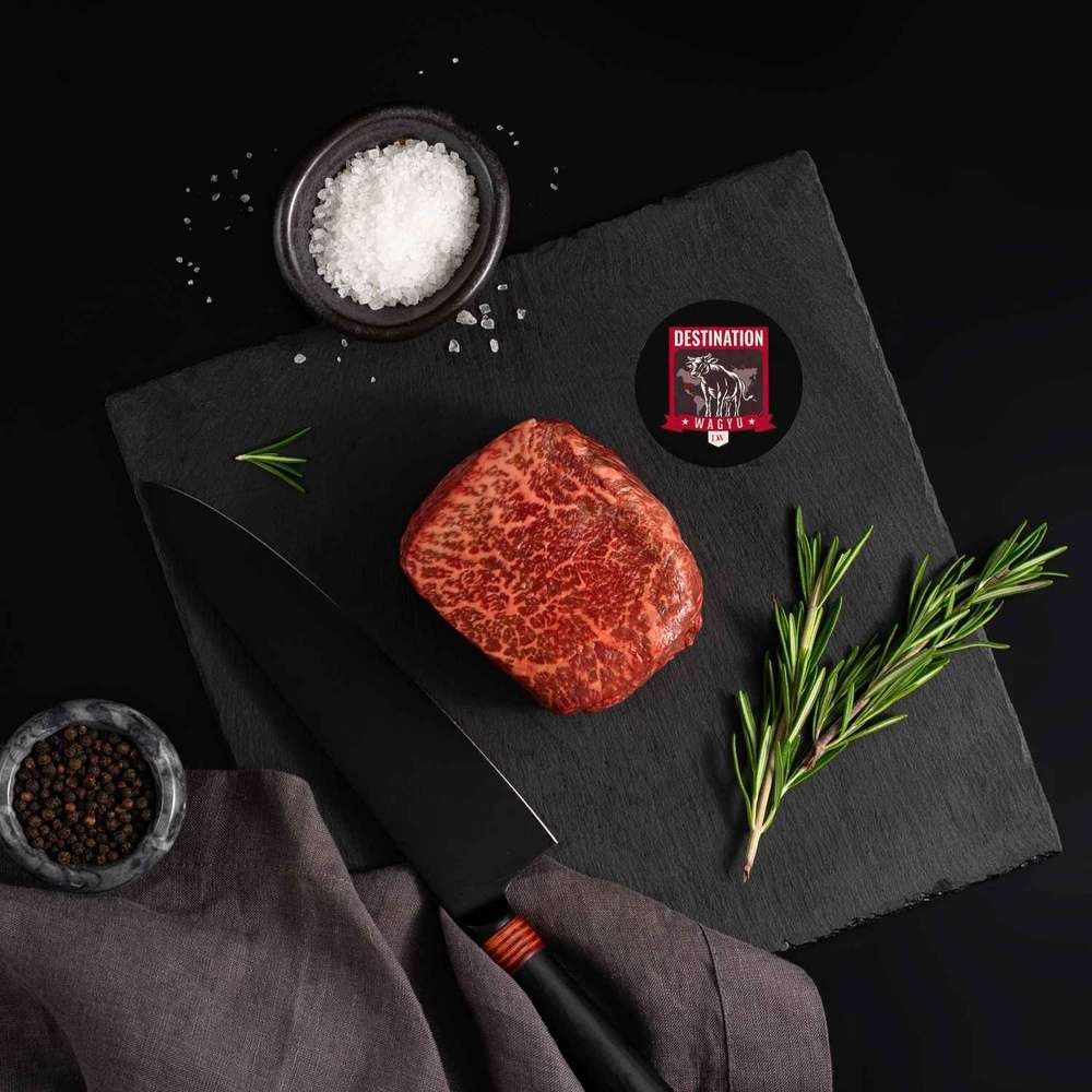 An Australian Wagyu Full Blood Top Sirloin Steak rests on a slate board with rosemary, a bowl of salt, a bowl of pepper, and a black knife.