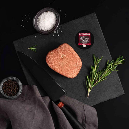 A Japanese A5 Wagyu Kobe Wine Filet Steak rests on a slate board with rosemary, a bowl of salt, a bowl of pepper, and a black knife.