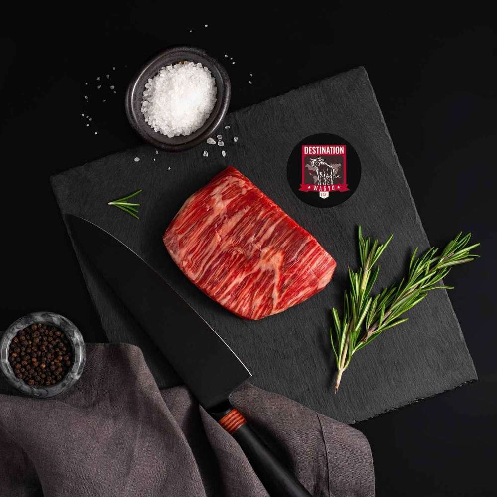 An Australian Wagyu Flank Steak rests on a slate board with rosemary, a bowl of salt, a bowl of pepper, and a black knife.