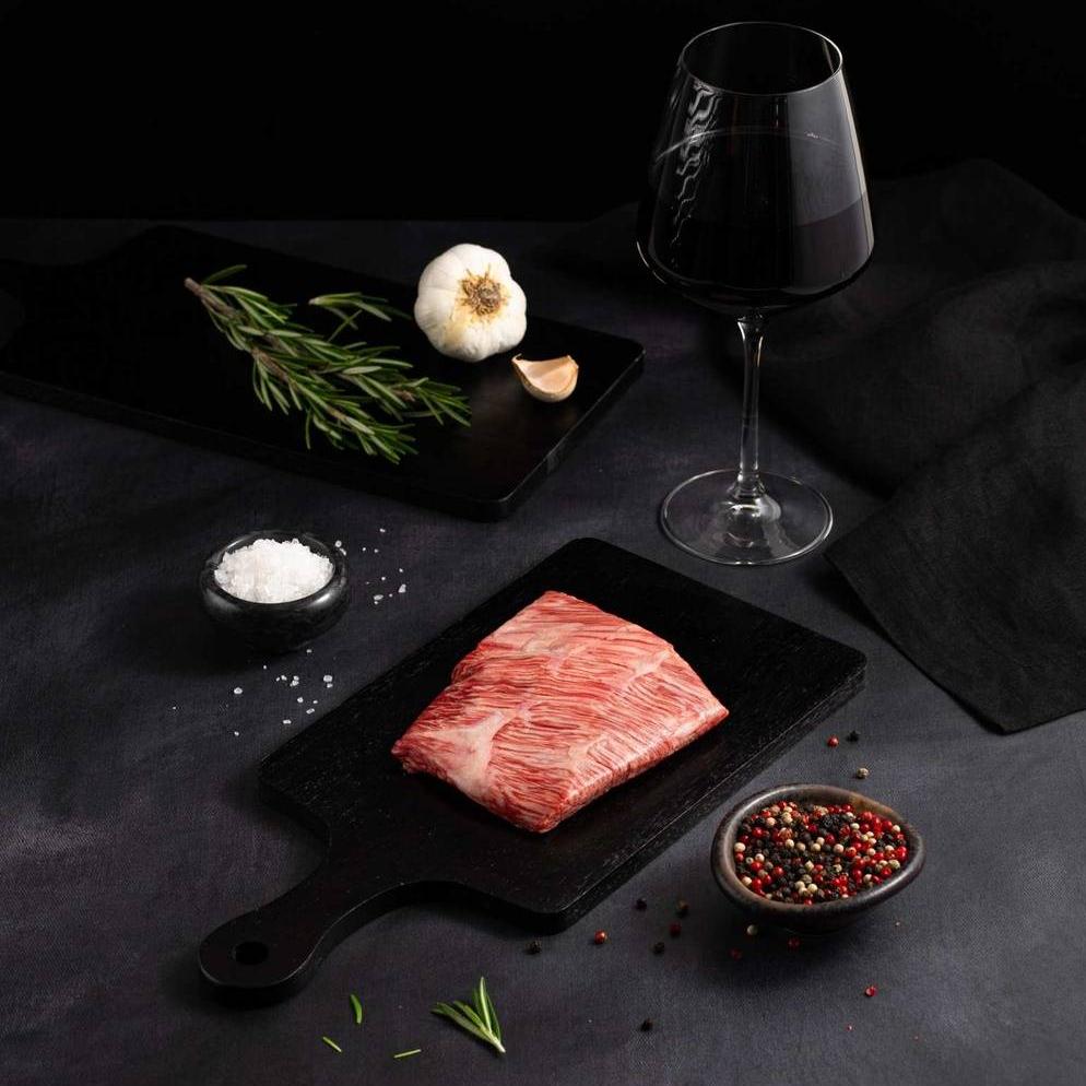 A Raw Japanese A5 Wagyu Rib Cap Steak rests on a black slate surrounded by salt, pepper, rosemary, garlic, and complemented by a glass of red wine on a dark surface.